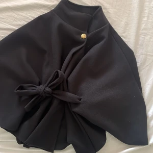 Svart poncho med bälte från Missay - Säljer en svart poncho från Missay med knytbälte i midjan och en dekorativ guldig knapp vid halsen. Oanvänd helt ny med prislapp 