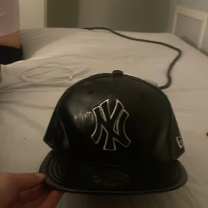 Svart New York Yankees keps i blankt material - Säljer en svart keps från New Era med New York Yankees-logga framtill i vitt. Kepsen har en blank finish och klassisk form med böjd skärm. NY-logga även bak och på sidan.