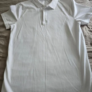 Vit pikétröja från Next School - Klassisk vit pikétröja från Next School i 100% bomull. Tröjan har korta ärmar, krage och knäppning med tre knappar framtill. Perfekt basic-plagg med enkel och stilren design.