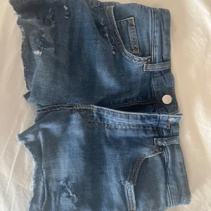 Lågmidjade Blå jeansshorts med fransar - Säljer ett par blå jeansshorts med fransiga benslut och slitna detaljer. Klassisk femficksmodell med knapp och dragkedja framtill. Perfekta för sommardagar och festivalhäng. Sitter som s