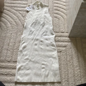 Vit ribbad ärmlös klänning från Bershka - Supersnygg vit ribbad klänning från Bershka i ärmlös modell med hög hals. Baksidan har en öppen rygg med en glittrig kedjedetalj. Perfekt för dig som vill sticka ut med en modern och trendig look. Aldrig använd