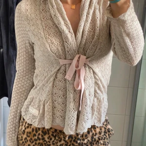 Beige stickad kofta med rosett från Odd Molly - Supersöt beige stickad kofta från Odd Molly med spetsmönster och ljusrosa band i midjan. Koftan har lång ärm och volangkant nertill, perfekt att slänga på över ett linne eller t-shirt.