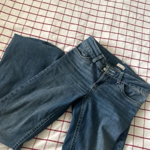Blå jeans med dekorficka - Säljer ett par klassiska blå jeans med utsvängda ben och snygga kontrastsömmar. På bakfickan finns en dekorativ detalj i form av ett mönster med små sydda  pärlor. Jeansen har två knappar i midjan och normal passform. Använt ett par få antal gånger. 