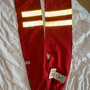 Röda GORE-TEX byxor med reflexdetaljer - Röda byxor med två breda reflexband runt låren och dragsko vid benslut. Tillverkade i slitstarkt GORE-TEX-material som står emot väta. Perfekta för utomhusaktiviteter där synlighet är viktigt.
