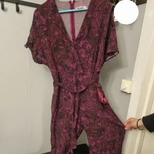 Mörklila omlottklänning med bälte - Säljer en mörklila byxdress  med kort ärm och bälte i midjan. Klänningen har ett paisleymönster i svart och lila, v-ringad hals och dragkedja bak. Perfekt för dig som vill ha något unikt och snyggt!