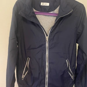Mörkblå vindjacka från H&M - Säljer en mörkblå vindjacka från H&M med vita dragkedjor framtill och vid fickorna. Jackan har lång ärm och en enkel, stilren design som passar perfekt till vår och höst.