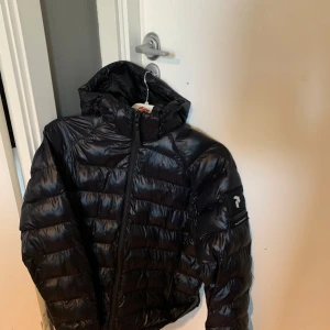 Svart pufferjacka från Peak Performance - Säljer en svart pufferjacka från Peak Performance med huva och dragkedja. Jackan har lång ärm, ribbade muddar och logotyp på ärmen. Perfekt för kyliga dagar och enkel att matcha med det mesta.