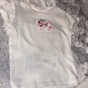 Vit t-shirt med rosett från H&M - Söt vit t-shirt från H&M med en rosa rosett och texten 'Kind Heart' framtill. T-shirten har korta ärmar och är gjord i mjuk bomull. Perfekt för en enkel och gullig stil. Den är också helt ny använt 0 gånger.