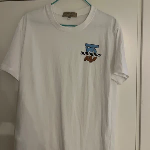Vit t-shirt från Burberry med logga - Säljer en vit t-shirt från Burberry med tryckt logga i blått och brunt på bröstet. Klassisk rund halsringning och normal passform. Perfekt för dig som gillar stilrena märkesplagg.