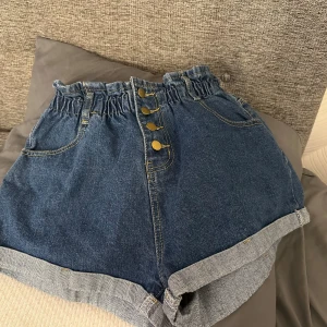 Blå högmidjade jeansshorts med knappar - Snygga blå jeansshorts med hög midja och fyra guldiga knappar framtill. Shortsen har papperspåse-midja och uppvikta benslut. Perfekta för varma dagar och enkel att matcha med olika toppar.