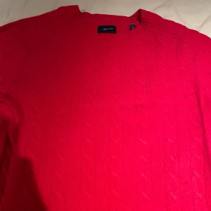 Röd kabelstickad tröja från GANT - Säljer en klassisk röd kabelstickad tröja från GANT med rund halsringning. Tröjan har diskret broderad logga på bröstet och ribbade muddar vid ärmslut och nederkant. Perfekt för lager-på-lager under kyliga dagar.