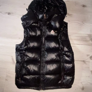 Svart dunväst från Moncler - Snygg svart dunväst från Moncler med glansig finish och huva. Västen har dragkedja framtill, två fickor med dragkedja och klassisk Moncler-logga på bröstet. Perfekt för lager-på-lager under kyliga dagar.