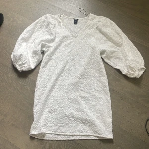 Vit omlottklänning med puffärmar - Superfin vit klänning med omlott-skärning och stora puffärmar. Klänningen har strukturmönster i tyget och v-ringad hals. Perfekt för somriga tillfällen och midsommar.