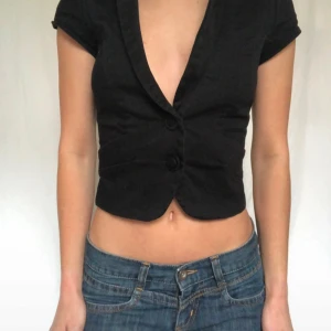 Svart croppad kavajliknande topp - Säljer en svart croppad topp med kavajkrage och korta ärmar. Toppen har två knappar framtill och djup v-ringning. Perfekt för en trendig och edgy look.