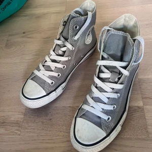 Grå Converse Chuck Taylor All Star - Klassiska höga Converse Chuck Taylor All Star i grått med vit tå och sula. Skorna har snörning och den ikoniska loggan på sidan.    . De har använts många gånger men är i bra skick!