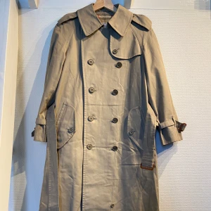 Vintage Burberrys trenchcoat - Tidlös klassisk beige trenchcoat från Burberrys med ikoniskt rutigt foder. Japansk produktion från 90-talet eller tidigare, se lapparna. Jackan har dubbelknäppning, krage, bälte i midjan och vid ärmslut samt stora fickor framtill. Passar som en storlek S (Herr). Mått: längd 110 cm, ärmlängd 52 cm, bredd 55 cm, axelbredd 45 cm. I utmärkt skick med tanke på att den är minst 30 är gammal. De enda defekterna är nästintill osynliga små fläckar, kan skicka närmare bild på dessa eftersom de inte syns.