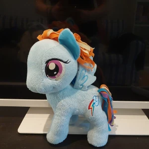 My Little Pony Rainbow Dash Mjukisdjur - Säljer en mjuk och färgglad Rainbow Dash-mjukisdjur från My Little Pony. Den är ljusblå med stora lila ögon, regnbågsfärgad man och svans i orange, blått och lila. På sidan finns en broderad blixt med moln.