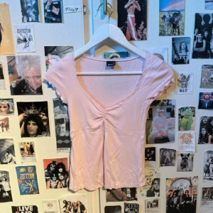 Gina topp - Söt ljusrosa topp med korta ärmar, v-ringning och rynkdetalj framtill. Toppen har en slits med spetskant som ger en extra feminin touch. Perfekt för dig som gillar romantisk stil. Aldrig använd