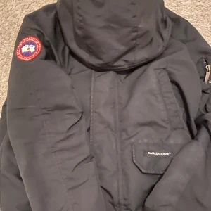 Svart vinterjacka från Canada Goose - Säljer en svart vinterjacka från Canada Goose med huva och klassisk logga på ärmen. Jackan har dragkedja och flera fickor, perfekt för kalla dagar. Stilig och funktionell design med tydlig Canada Goose-märkning.
