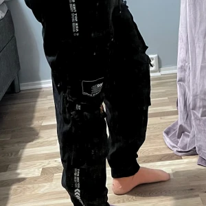 Svarta cargopants med tryck - Svarta cargopants med flera fickor och vita samt orange tryckdetaljer på benen. Byxorna har en loose passform och dragsko i midjan. Perfekta för en avslappnad streetwear-look.