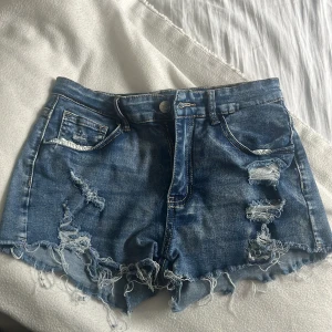Blå jeansshorts med fransar och slitningar - Snygga blå jeansshorts med råa fransar nertill och flera slitningar både fram och bak. Klassisk femficksmodell med knapp och dragkedja. Perfekta för sommaren och ger en cool, avslappnad look.