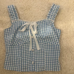 Blåvit rutig linnetopp med rosett och knappar - Supersöt blåvit rutig linnetopp med breda axelband, dekorativ rosett framtill och pärlknappar. Toppen har spetsdetaljer vid urringningen och smockad rygg för extra komfort. Perfekt för varma dagar!