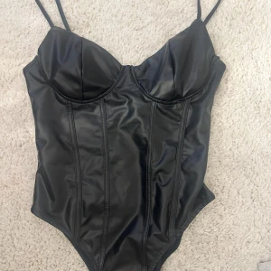 Svart korsett-topp med smala axelband - Säljer en svart korsett-topp med smala axelband och markerade kupor. Toppens material har en glansig finish som ger en cool look. Perfekt att styla till olika outfits för en edgy vibe.