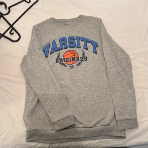 Grå sweatshirt med baskettryck från Primark - Säljer en grå sweatshirt från Primark med blå text 'VARSITY' och en orange basketboll på bröstet. Tröjan har långa ärmar, rund hals och ribbade muddar. Perfekt för en sportig look.