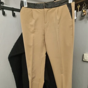 Beige kostymbyxor med svarta detaljer från Reserved - Snygga beige kostymbyxor från Reserved med svarta skinn detaljer i midjan och vid fickorna. Byxorna har en klassisk rak passform och är perfekta för en stilren look. Tillverkade i ett mjukt material som känns bekvämt hela dagen.