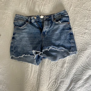 Blå jeansshorts med fransar - Säljer ett par klassiska blå jeansshorts med råa fransade kanter. Shortsen har fem fickor och stängs med knapp och dragkedja. Perfekta för varma dagar och enkel att matcha med olika toppar.