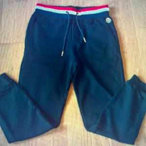 Svarta mjukisbyxor från Moncler med röd och vit midjerand - Svarta mjukisbyxor från Moncler med elastisk midja i rött, vitt och blått samt dragsko. Byxorna har Moncler-logga på höften och är perfekta för en avslappnad stil.