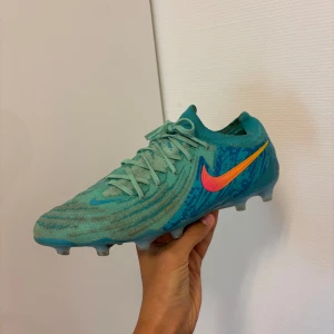 Turkosa Nike fotbollsskor - Säljer ett par turkosa Nike fotbollsskor med orange och rosa swoosh. Skorna har snörning och är designade för spel på gräs. Mönstrad ovandel och rund tå ger bra grepp och kontroll på planen. Ingen box inga sulor kommer med. 
