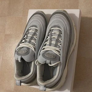 Nike Air Max 97 i grått - Säljer ett par Nike Air Max 97 i ljusgrått med vita detaljer. Skorna har den klassiska vågiga designen, snörning och en synlig Air Max-sula för extra komfort. Perfekta för dig som gillar stilrena sneakers med retrovibbar.