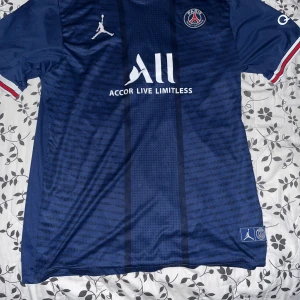 PSG hemmatröja Mbappé 7 - Mörkblå PSG fotbollströja med Mbappé 7-tryck på ryggen. Tröjan har vita och röda detaljer på ärmsluten, Jordan-logga på bröstet och klubbmärke. Tillverkad i lätt och ventilerande material.