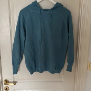 Blå hoodie med huva - Säljer en enkel blå hoodie med huva och dragsnören. Tröjan har långa ärmar och ribbade muddar vid ärmslut och nederkant. Perfekt för en avslappnad stil.
