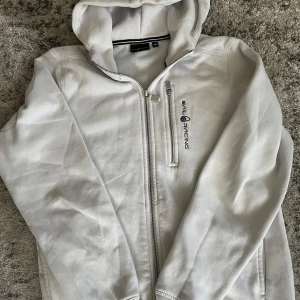 Vit hoodiejacka från Sail Racing - Vit hoodiejacka från Sail Racing med dragkedja framtill och logga på bröstet. Jackan har huva och en bröstficka med dragkedja. Perfekt för dig som gillar sportig stil och vill ha något enkelt men snyggt.