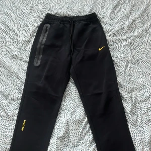 Svarta mjukisbyxor från Nike NOCTA - Svarta mjukisbyxor från Nike NOCTA med gul logga och detaljer. Byxorna har resår i midjan och raka ben. Snygga svarta ränder längs ena benet och NOCTA-tryck i gult. Perfekta för en sportig och trendig look.