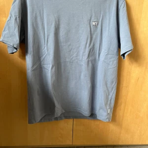 Blå t-shirt med WB-logga - Säljer en stilren blå t-shirt från WB med liten broderad logga på bröstet. T-shirten har normal passform och är tillverkad i mjuk bomull. Perfekt för en avslappnad look.