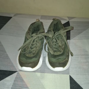 Olivgröna chunky sneakers - Säljer ett par olivgröna chunky sneakers med vit sula. Skorna har snörning och är tillverkade i syntetmaterial med meshdetaljer för extra andningsförmåga. Perfekta för dig som gillar en sportig och trendig stil.