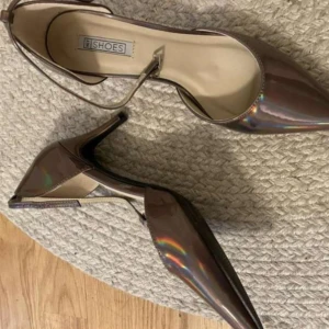 Klackar från Nelly - Säljer ett par glansiga bronsfärgade klackskor från Nelly med spetsig tå och smal klack. Skorna har en elegant ankelrem och en holografisk effekt som lyfter fin outfit💗