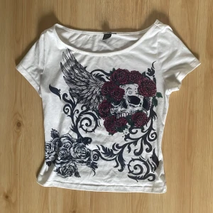 Vit croppad t-shirt med döskalle och rosor - Säljer en vit croppad t-shirt från New Yorker med stort tryck av döskalle, rosor och vingar i svart och rött. T-shirten har korta ärmar och en bred, något båtringad hals. Perfekt för dig som gillar edgy stil!