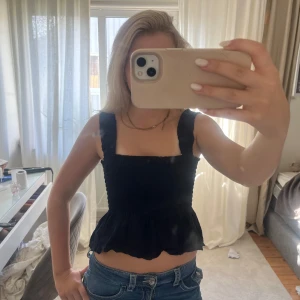 Svart croppad linnetopp med volang - Säljer en svart croppad linnetopp med breda axelband och volang nertill. Toppen har smockad struktur och fyrkantig ringning, perfekt för sommaren.