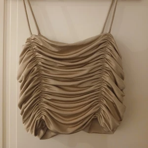 Beige omlott topp med rynk från AMISU - Säljer en beige omlott topp från AMISU med smala axelband och snygga rynkade detaljer framtill. Toppen har en glansig finish och är perfekt för att skapa en trendig look.