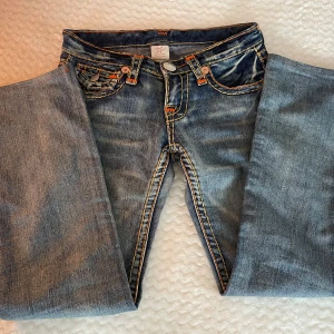 Blå jeansbyxor från True Religion - Säljer ett par klassiska blå jeans från True Religion med stora bakfickor med detaljer. Sjukt snygga och är vintage så går inte att få tag på längre. Gylfen är dock lite trasig då gylfen ramlat av men går enkelt att sy dit en ny eller med hjälp av ett gem!