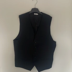 Svart kostymväst från Filippa K - Stilren svart kostymväst från Filippa K med klassisk passform. Västen har knäppning framtill med fem knappar, tre fickor och justerbart band i ryggen. Perfekt för en dressad look.