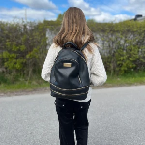 Svart ryggsäck från River Island - Säljer en snygg svart ryggsäck från River Island med gulddetaljer och flera dragkedjefack. Väskan är tillverkad i ett strukturerat syntetmaterial och har en stilren design med en guldig logoplatta framtill. Perfekt för att bära med sig allt man behöver till skolan eller stan.
