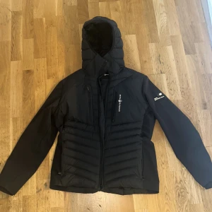 Svart dunjacka från Sail Racing - Säljer en svart dunjacka från Sail Racing, modell Antarctica Expedition. Jackan har huva, dragkedja framtill och flera fickor. Perfekt för kalla dagar och har en sportig look med logga på ärmen.