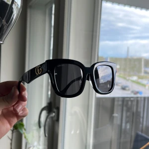 Svarta solglasögon från Gucci - Stilrena svarta solglasögon från Gucci med fyrkantig form och breda skalmar. GG-logga i guld på sidorna ger en lyxig känsla. Glasögonen har mörka linser och är perfekta för att lyfta din look.