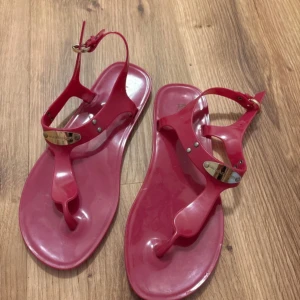 Rosa sandaler från Michael Kors - Säljer ett par snygga rosa sandaler från Michael Kors i syntetmaterial. De har en tårem och justerbart spänne vid ankeln samt guldfärgade metalldetaljer framtill. Perfekta för varma dagar!