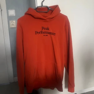Orange hoodie från Peak Performance - Säljer en orange hoodie från Peak Performance med svart logga tryckt på bröstet. Tröjan har huva och en stor magficka framtill. Perfekt för dig som gillar en enkel och stilren look.